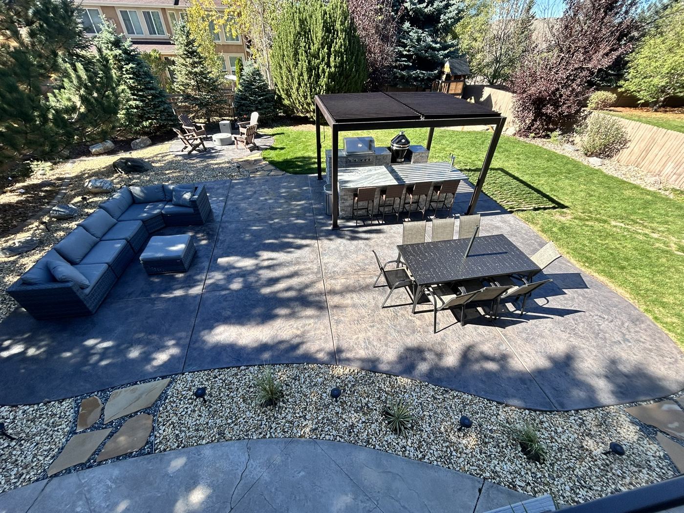 After: the same backyard with a stamped concrete patio, outdoor sectional, dining table, and a louvered pergola over an outdoor kitchen and bar.