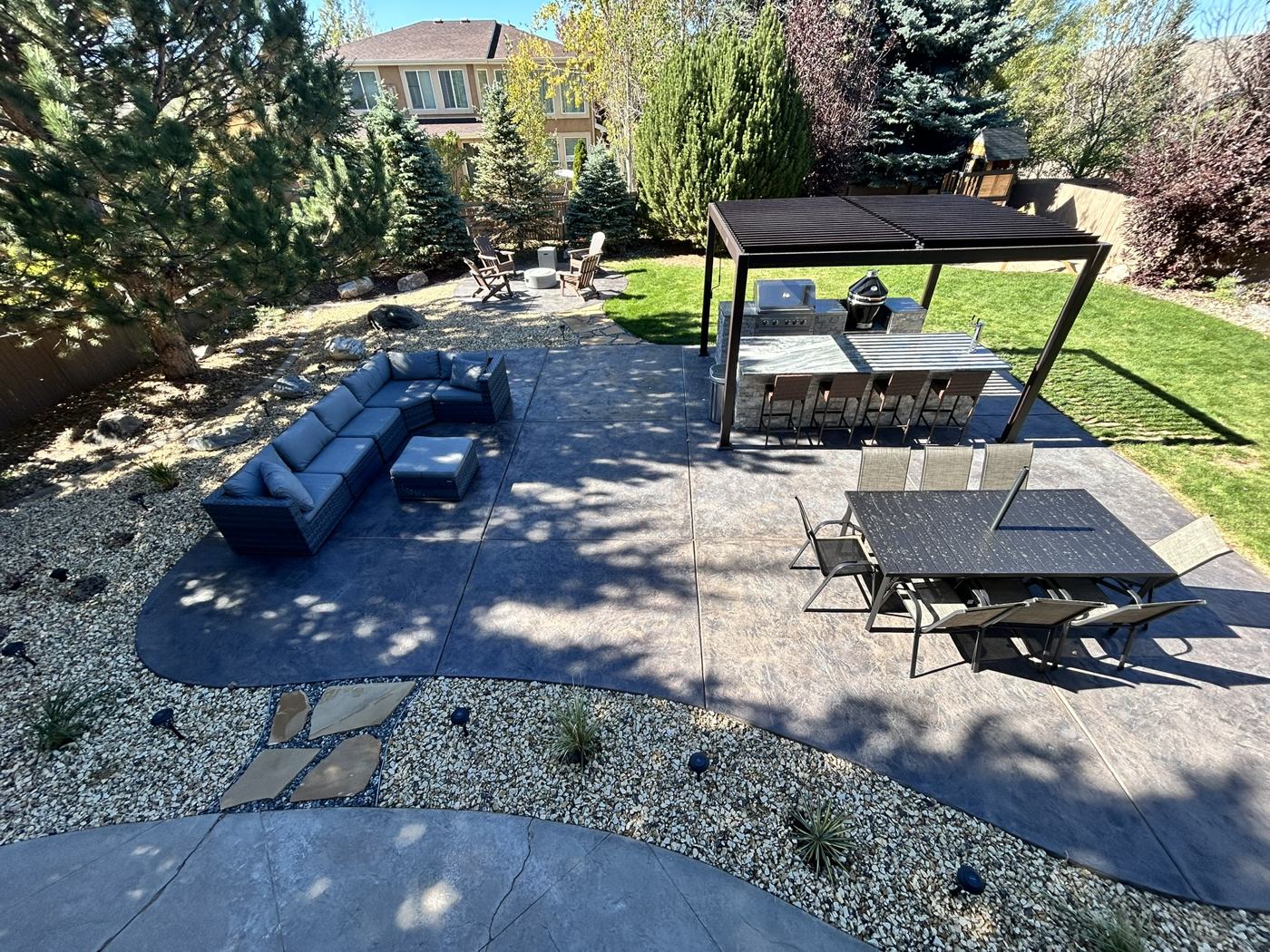 A finished backyard with a stamped concrete patio, louvered pergola, outdoor kitchen, seating areas and rock landscaping, seen from above.