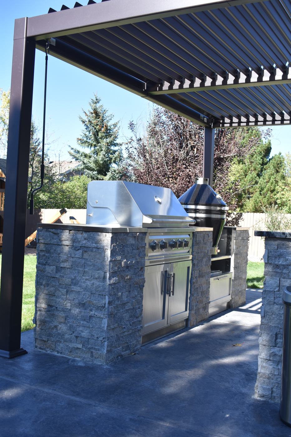 A stone-clad grill station with stainless doors and a kamado smoker under a louvered pergola.
